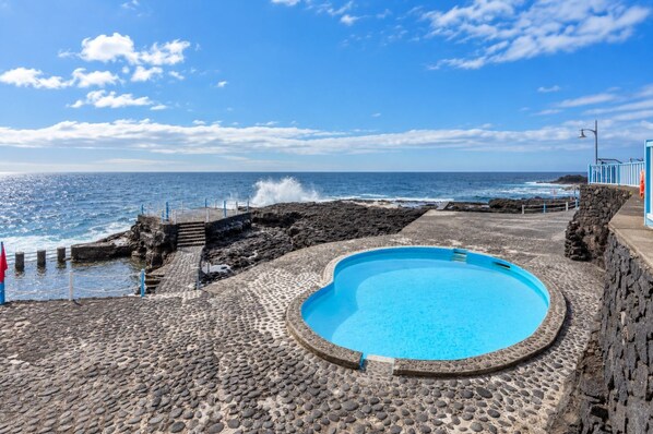 A pool with a stone pathway leading to the sea, a stone wall, and a clear view of the ocean.