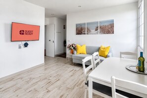 A modern living room with a flat-screen TV, a sofa with yellow cushions, a dining table with chairs, and a painting on the wall.