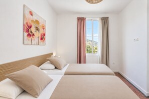 A hotel room with two beds, a window with curtains, a painting on the wall, and a view of the outside.