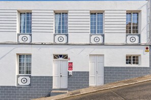 Un edificio con fachada simétrica, que presenta una puerta central con un letrero, varias ventanas con contraventanas y un ventanal con vista al interior.