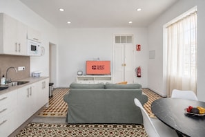 Un espacio habitable compacto con kitchenette, un sofá gris, una mesa de comedor con una bandeja de fruta y una pantalla de televisión que muestra 'GUEST READY'.