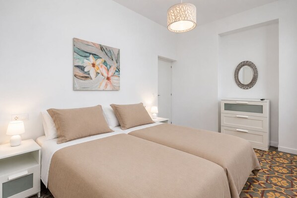 Une chambre à coucher avec un lit double, des tables de chevet avec des lampes, une commode, un miroir et un tableau représentant des motifs floraux accroché au mur.