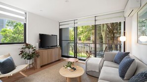 Uma sala de estar moderna com televisão de tela plana, sofá, poltrona, mesa redonda pequena e varanda com vista para árvores.