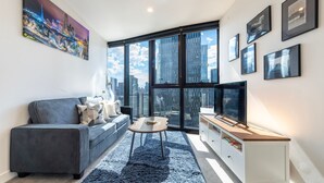 A modern living room with a sofa, coffee table, and TV, large windows with a city view, and framed pictures on the wall. - Southbank Skyline Suite - City Views & Prime Location (Southbank)