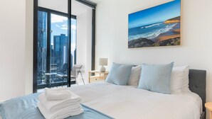 A hotel room with a bed, a view of the cityscape through large windows, a painting of a beach, and a small table with a lamp. - Southbank Skyline Suite - City Views & Prime Location (Southbank)