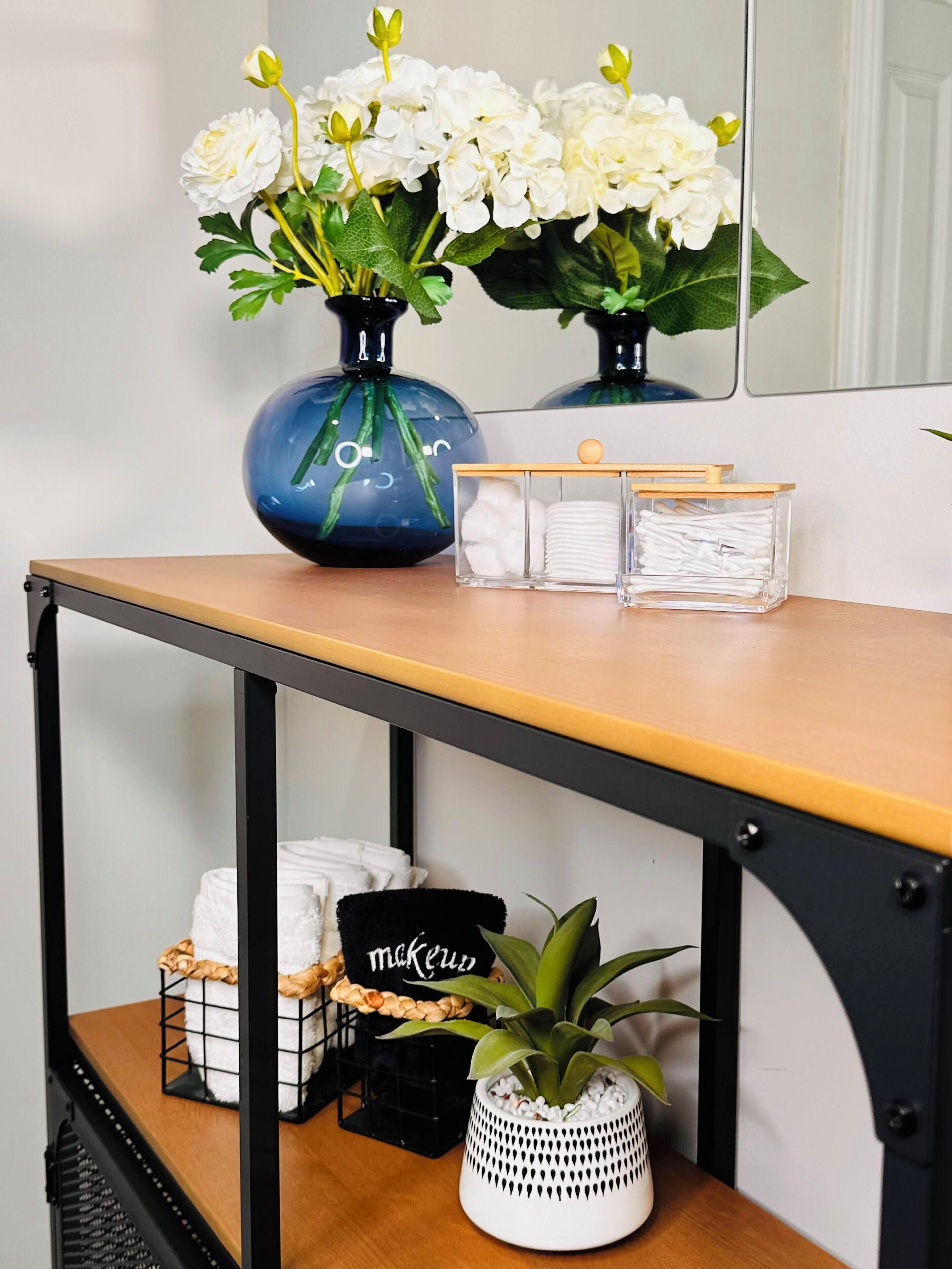 Una mesa de madera con un jarrón de flores blancas, una pequeña planta en maceta y una cesta negra con un letrero.