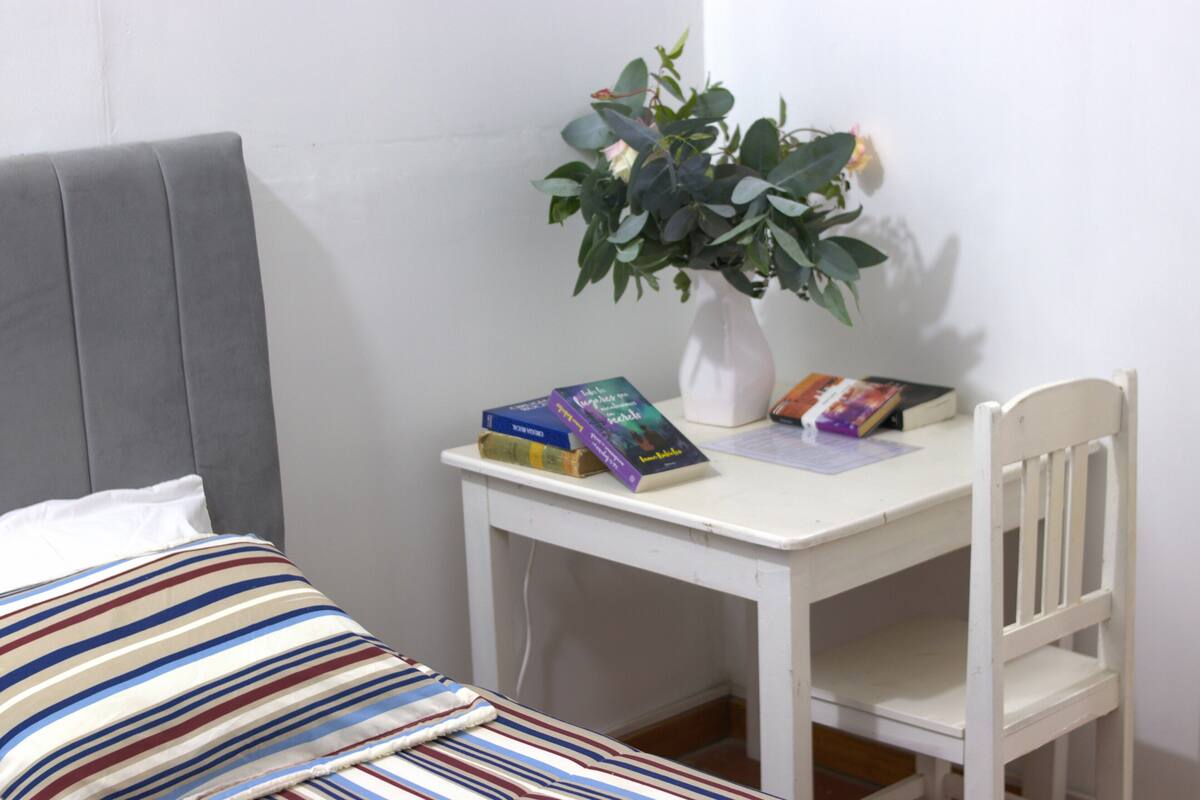 Ein Bett mit einem gestreiften Kopfkissen, ein weißer Schreibtisch mit einer Pflanzvase und Bücher.