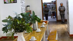 Un comedor con una larga mesa de madera, tazas blancas, un termo plateado y una planta en maceta.