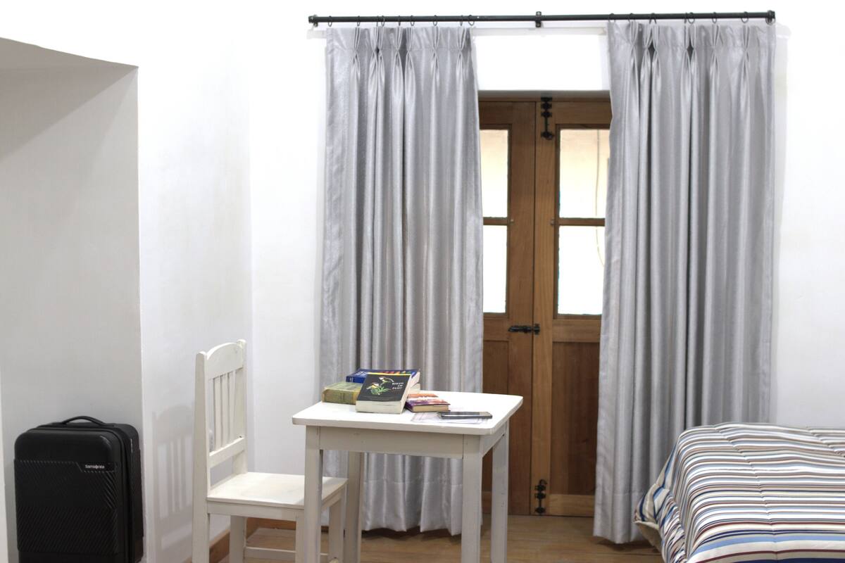 Ein Schlafzimmer mit einem Bett, einem kleinen Tisch mit Büchern, einem Stuhl, einem Koffer und einer Tür mit Vorhängen.