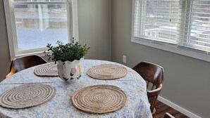 A dining area with a round table covered by a floral tablecloth, surrounded by wooden chairs with cushioned seats. - The Nest at Black Hall (Old Lyme)