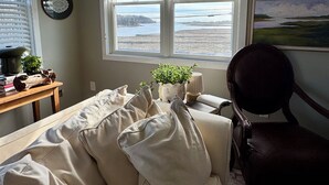 A room with a window, a couch, a chair, a table with a plant, and a wall with a painting. - The Nest at Black Hall (Old Lyme)