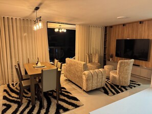Una sala de estar moderna con comedor, un conjunto de sofás, un televisor y un ventanal con cortinas.