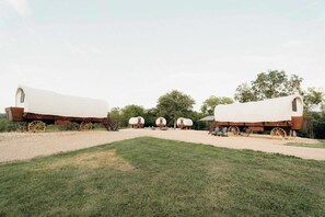 A row of covered wagons with wooden wheels, arranged in a line on a gravel path with grass in the foreground. - Glamping Wagon with Lake access + Golf carts (Branson)
