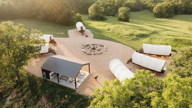 A rustic outdoor setting with covered seating areas, a central fire pit, and a small building with a sloped roof. - Cozy Couples Wagon | Lake Access + King bed (Branson)