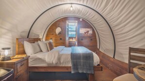 A cozy tented room with a wooden bed, a nightstand, and a window with a view. - Cozy Couples Wagon | Lake Access + King bed (Branson)