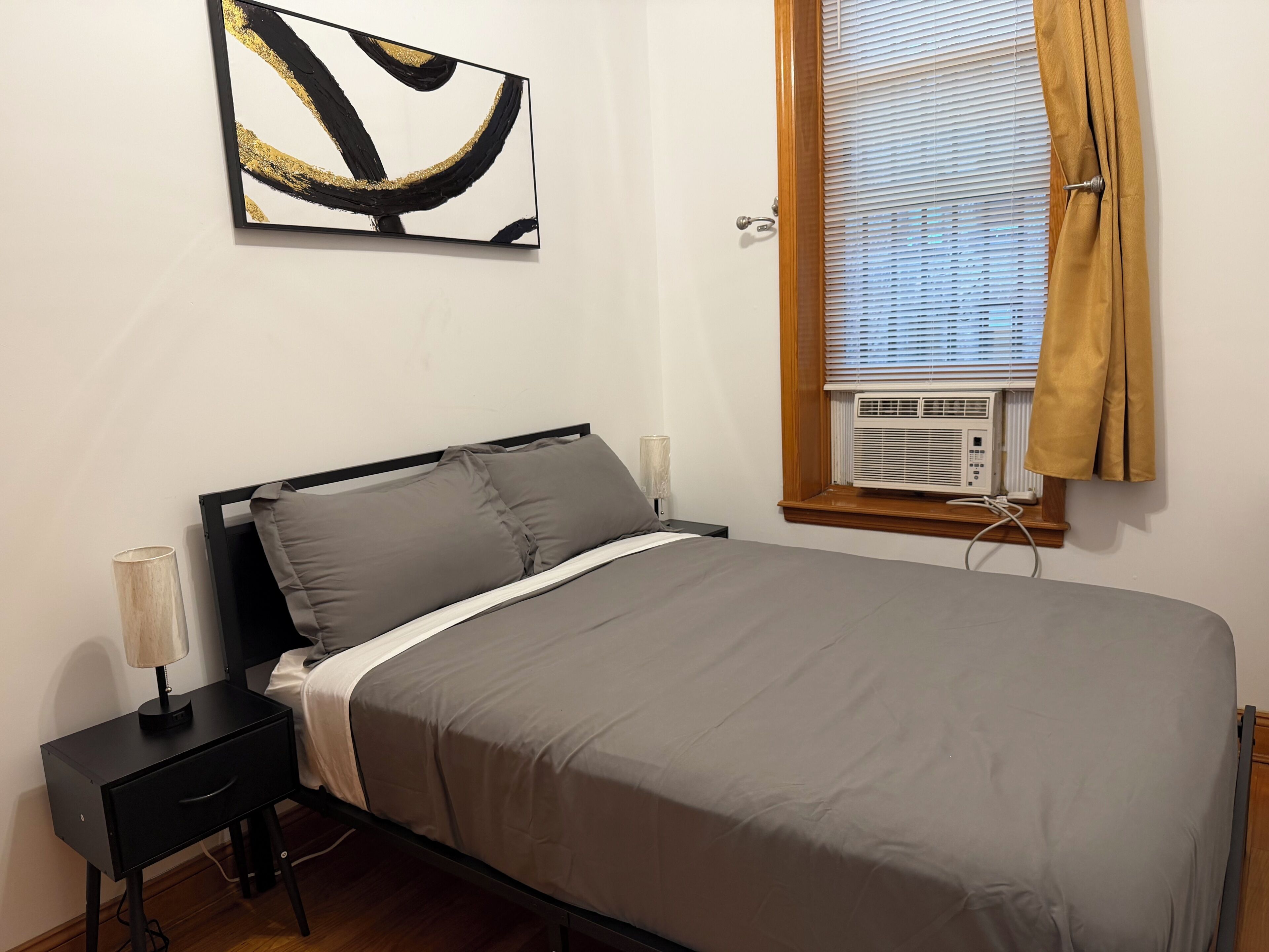 Une chambre à coucher comprenant un lit, des tables de chevet, une lampe, une fenêtre avec des stores et un climatiseur fixé au mur.