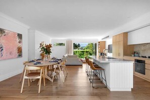 A modern kitchen with a central island, wooden dining table, and chairs, with a view of greenery outside. - Hidden Oasis – A Private Tropical Retreat in the Heart of Berry (Shoalhaven City Council)