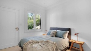 Ein Schlafzimmer mit einem Bett, einem Fenster mit Jalousien, einem Nachttisch mit Lampe und einer Tür.