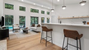 A modern kitchen with a white countertop, bar stools, and a dining area with a sofa and coffee table. - New Listing! Luxury Pondside Cabin | Hot Tub (McArthur)