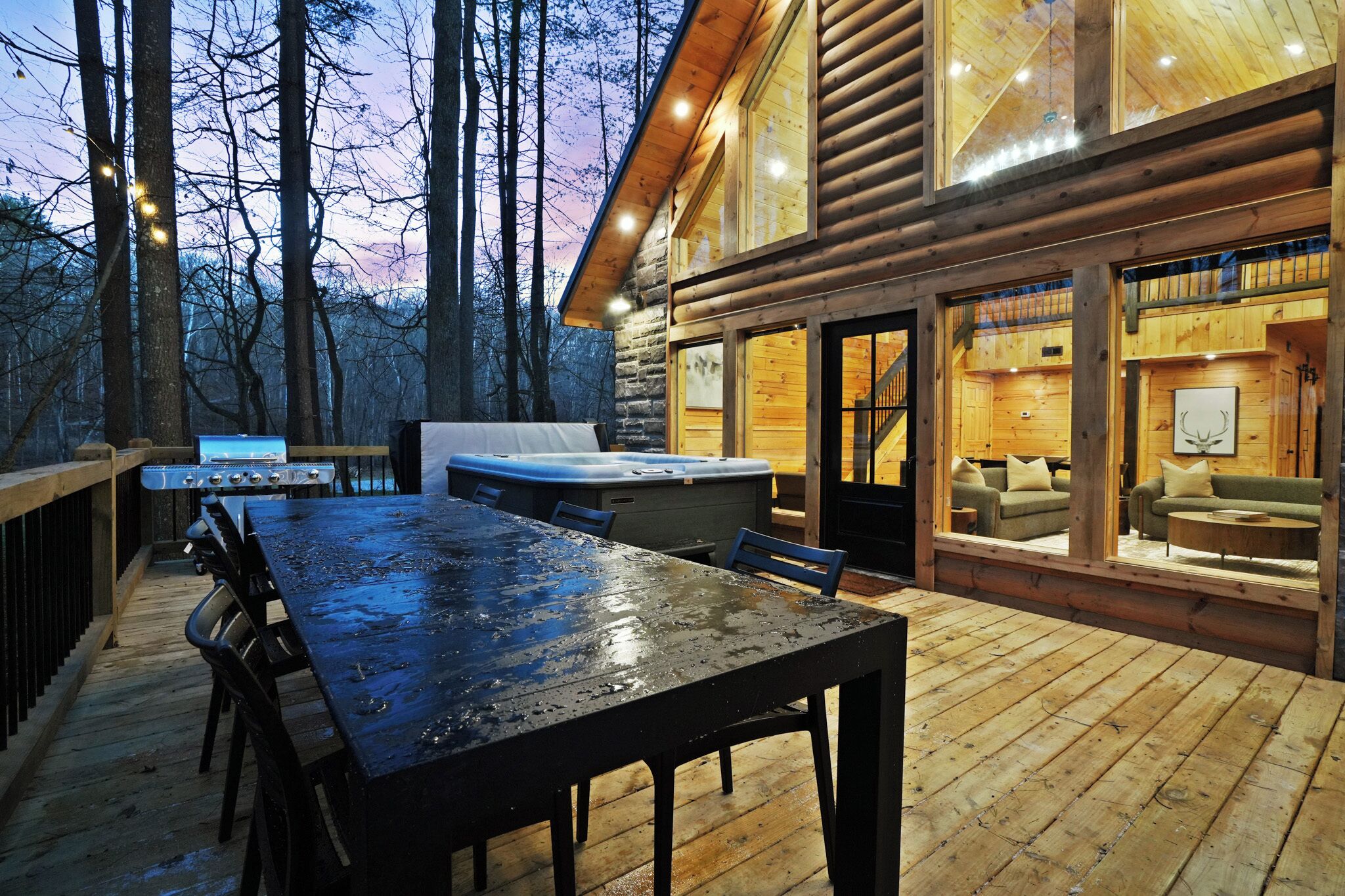 A wooden deck with a table and chairs, a hot tub, and a sofa area with a view of the forest.