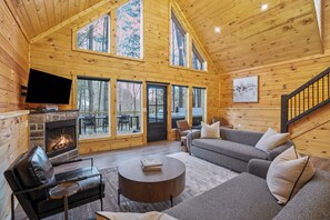 A log cabin interior with a fireplace, sofa, television, and large windows. - New! Luxury Log Cabin | Hot Tub | Dog Friendly (Creola)