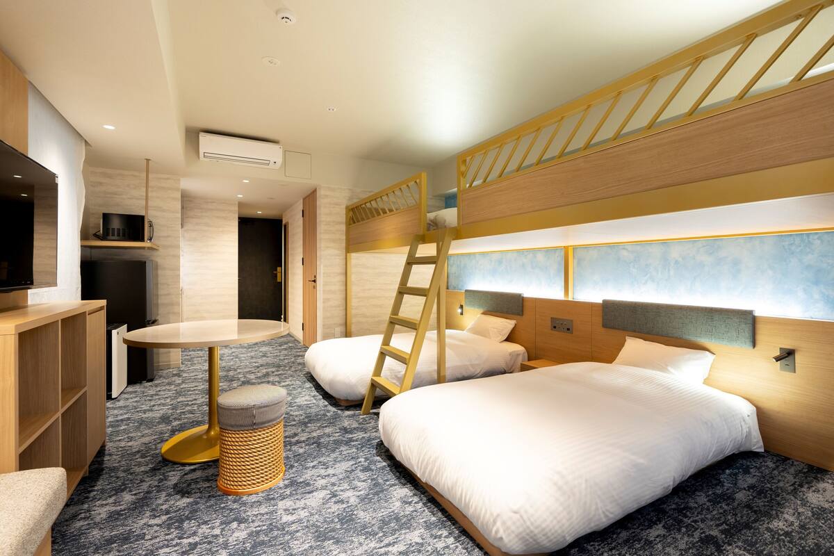 Ein Hotelzimmer mit Etagenbetten, einem kleinen Tisch, einem Stuhl, einem Fernseher und einer Küchenzeile.