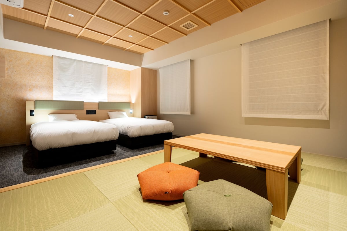 Ein Hotelzimmer mit zwei Betten, einem Holztisch, Tatami-Matten und einem großen Fenster mit Jalousien.