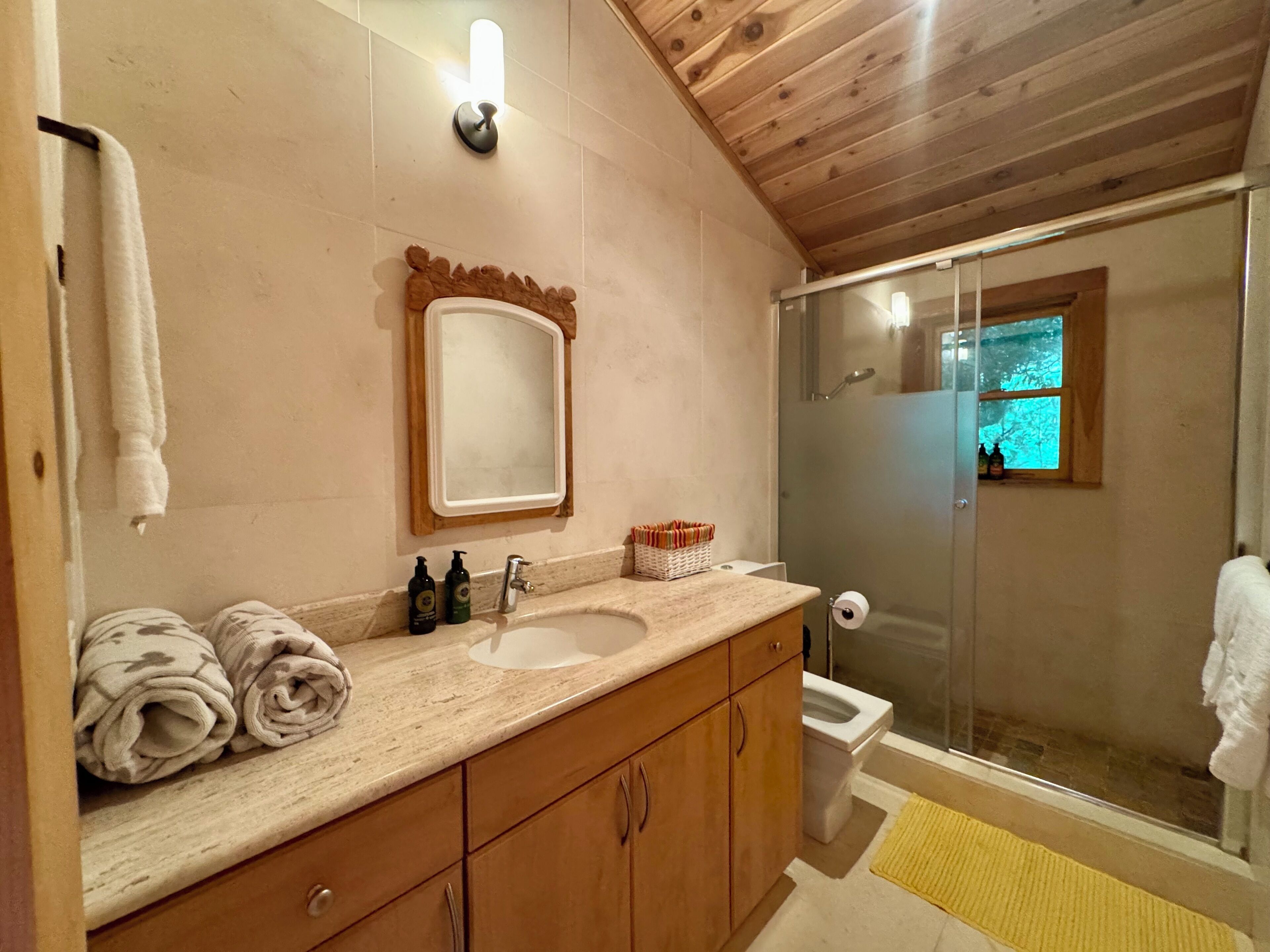 A bathroom with a wooden vanity, a sink, a mirror, a glass shower enclosure, and a window.
