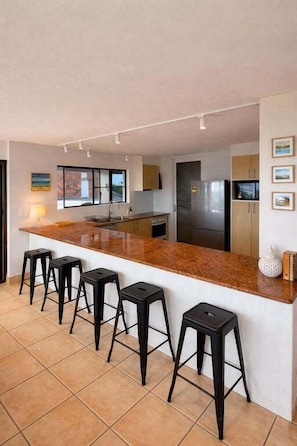 A modern kitchen with a central island, bar stools, a refrigerator, and a microwave. - Blue Sea View - Mooloolaba (Mooloolaba)