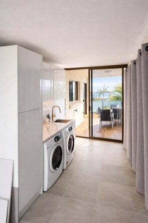 A modern kitchen with a built-in washing machine, a large window, and a view of a balcony with outdoor furniture. - Blue Sea View - Mooloolaba (Mooloolaba)