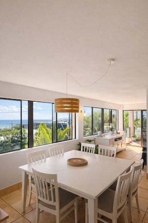 A spacious dining area with a white table and chairs, large windows offering a view of the outdoors, and a hanging light fixture. - Blue Sea View - Mooloolaba (Mooloolaba)