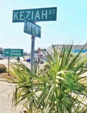 Panneaux de signalisation indiquant les rues KEZIAH ST et E. OAK ISLAND DR, un panneau directionnel indiquant la direction de BEACH STRAND, et un palmier au premier plan.