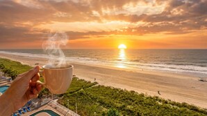 Une main tenant une tasse de laquelle s’échappe de la vapeur, avec en arrière-plan une plage et un coucher de soleil.
