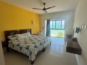 A bedroom with a bed, a ceiling fan, a window with a view of the beach, and a small desk with a lamp. - Complete Beachfront Villa with Pool | 5 Bedrooms | 8 Baths (Frente al mar)