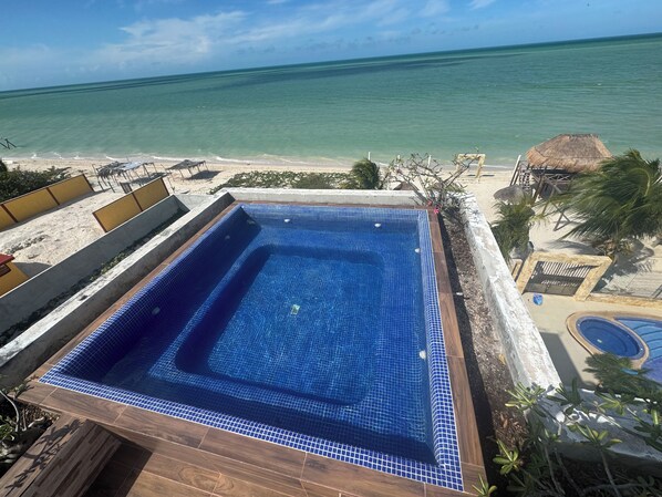 A swimming pool with a view of the ocean, surrounded by a wooden deck and a sandy beach area.