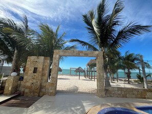 A stone archway leading to a beach with palm trees, a pool, and a thatched-roof structure in the background. - Complete Beachfront Villa with Pool | 5 Bedrooms | 8 Baths (Frente al mar)