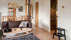 Ein Wohnzimmer mit einer Couch, einem Couchtisch und einem kleinen schwarzen Hocker. Eine Holztreppe, eine Tür zu einer Küche und ein Gemälde an der Wand sind ebenfalls zu sehen.