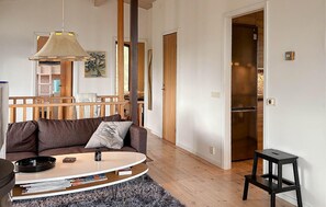 Un salon comprenant un canapé, une table basse et un petit tabouret noir. On y trouve un escalier en bois, une porte donnant sur une cuisine et un tableau accroché au mur.