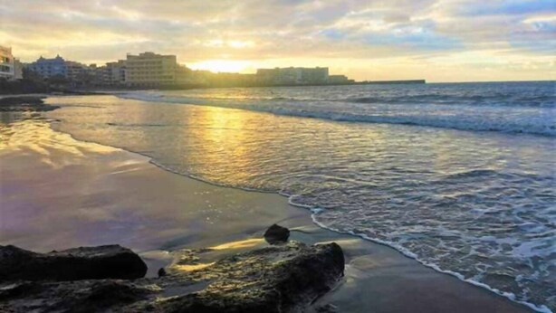 Uma praia com ondas, um pôr do sol e edifícios ao fundo.