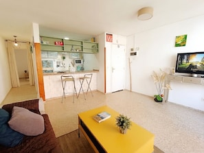A living room with a sofa, a yellow coffee table, and a TV. There is a kitchen area with a microwave and a sink. - Tenerife Sur El Médano Vacaciones - 3a (Medano, El)