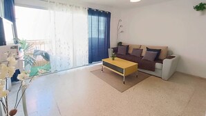 Sala de estar com sofá, mesa de centro, planta e vista pela janela com cortinas.