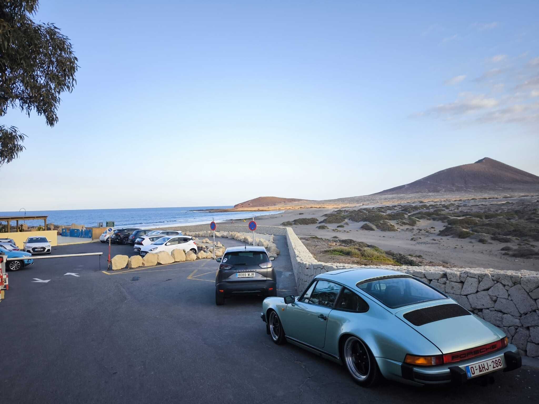 Un estacionamiento con coches, una playa y una montaña al fondo.