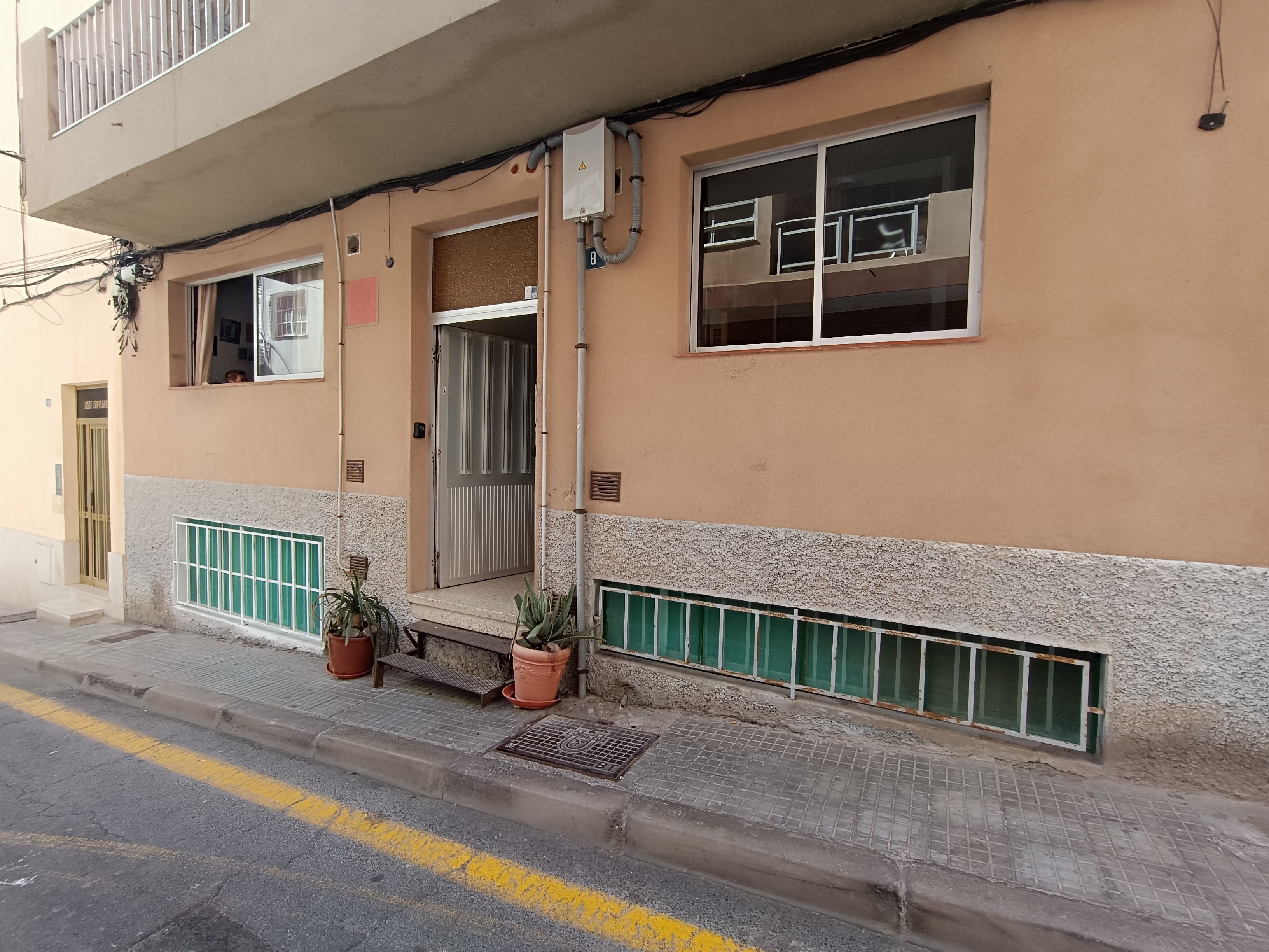 Un edificio con entrada protegida por una reja, una ventana con baranda metálica y una planta en maceta en la acera.