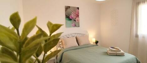 A bedroom with a bed, a bedside table, a lamp, a wall-mounted picture, and a potted plant.