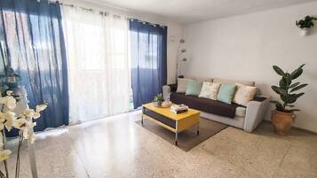 Una sala de estar con un sofá, una mesa de centro, una planta en maceta y un ventanal con cortinas.