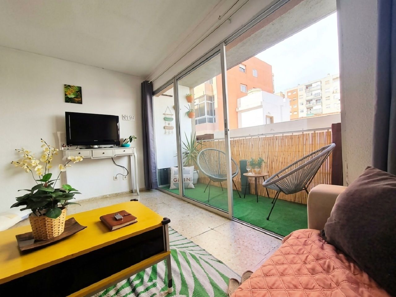 Una sala de estar con televisor, una mesa amarilla, un sofá y un ventanal corredizo que da a un balcón con una silla y una mesita.