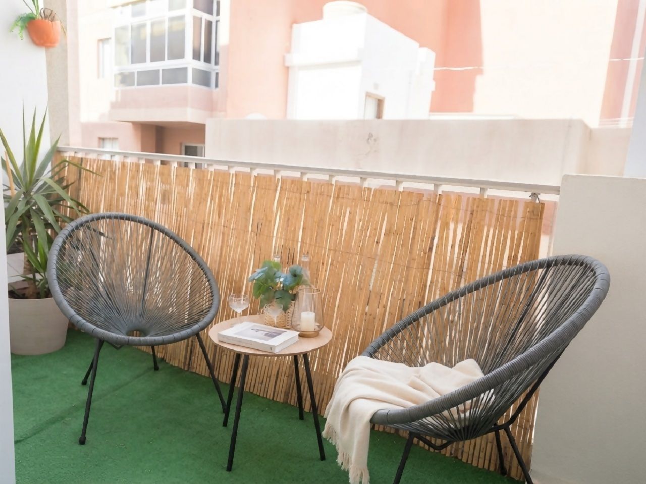 Een balkon met twee rieten stoelen, een klein rond tafeltje met een boek en kaarsen, en een bloempot.