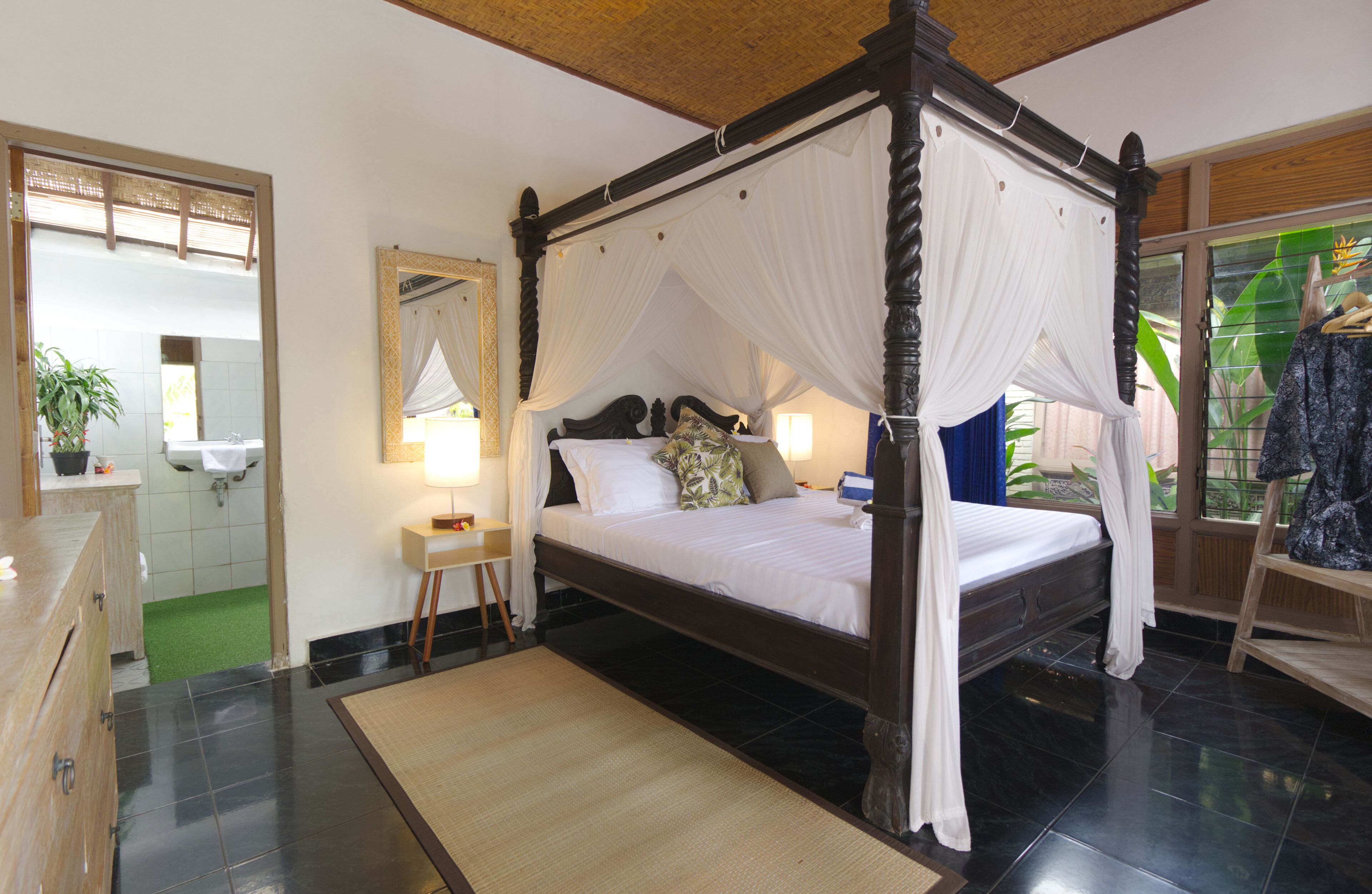 A four-poster bed with white bedding, a wooden nightstand with a lamp, a mirror, and a view of a green outdoor area.