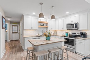 A modern kitchen with white cabinetry, a central island with stools, pendant lighting, and stainless steel appliances. - Gratitude 𓇼 Pool + Golf Cart + 0.7 mi to Beach (Oak Island)