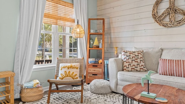 Ein gemütliches Wohnzimmer mit Sofa, Sessel, Holzregal und einem großen Fenster mit Vorhängen.
